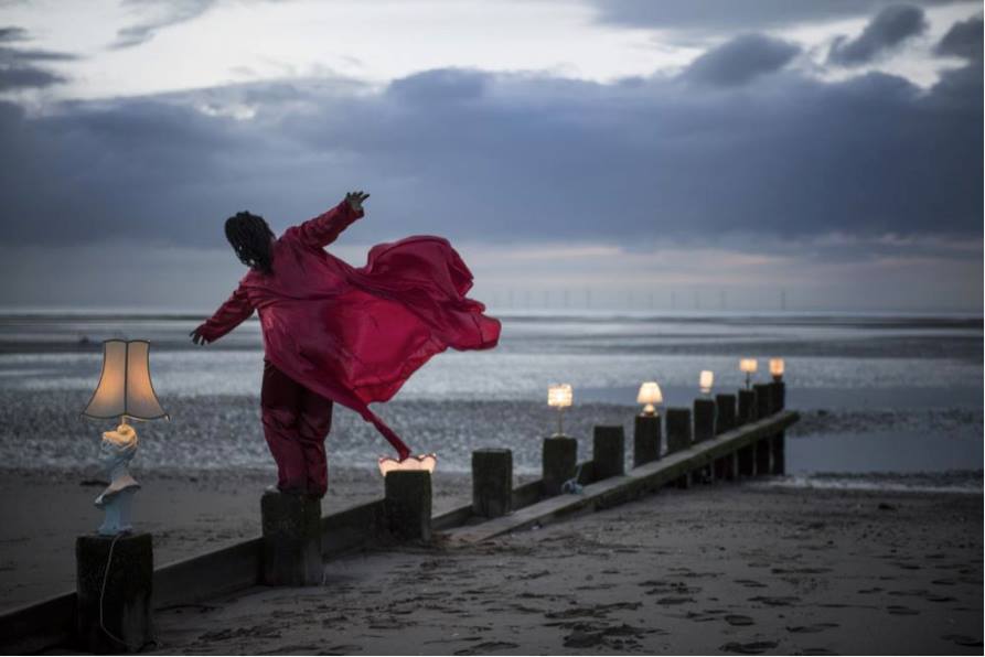 june stands on wave breakers on a beach, red gown flowing behind her in the wind, she stretches her arms out as if to fly 