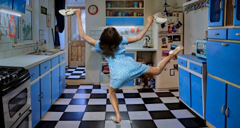 Roots image, dancer in a blue kitchen with back to the camera and legs in the air holding shoes.