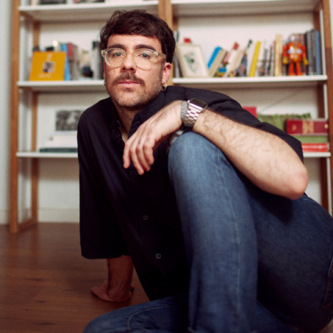 Marcos Morau sits on the floor in front of a trendy book case, he is white with a moustache and wears glasses