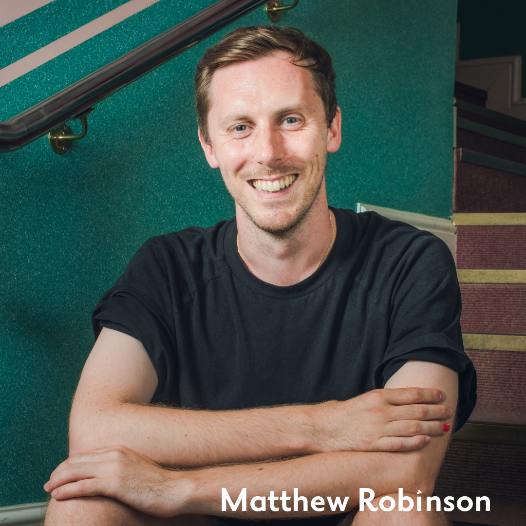 matthew robinson sits on some green and pink stairs smiling warmly, his arms crossed on his bare knees