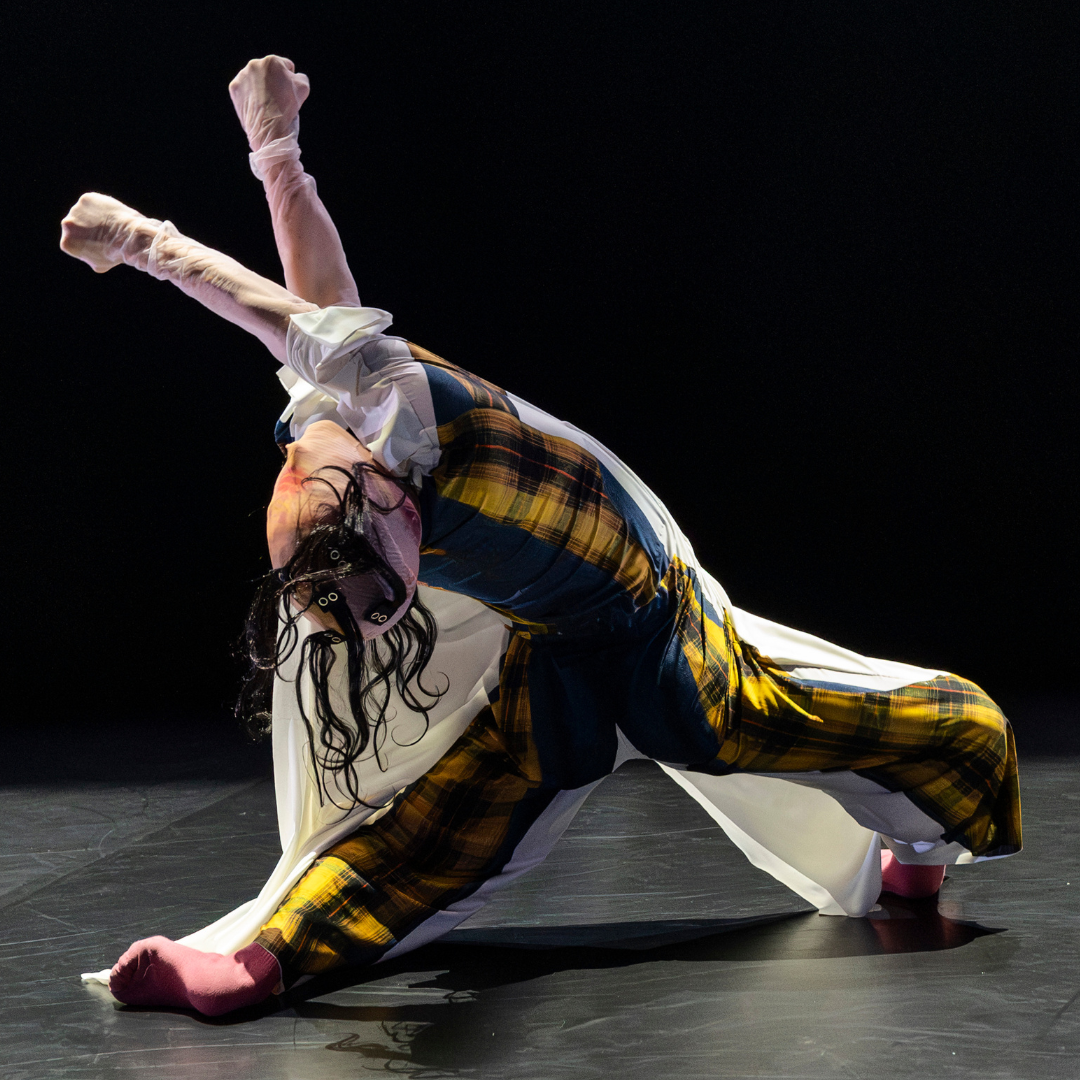 dancer leaning right back arms in air, in white costumes printed with 2d images of clothing and strange masks that distort some of their faces