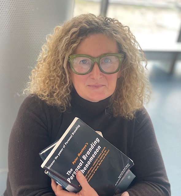 Joanna has blonde very curly hair and thick rimmed glasses, she's holding copies of a book