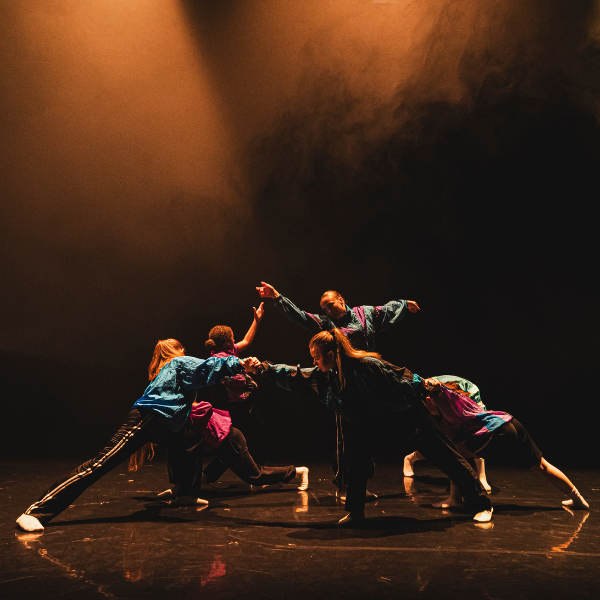 dancers in jewel toned vintage tracksuits lean towards one another under warm lighitng creating a traingle of arms and legs