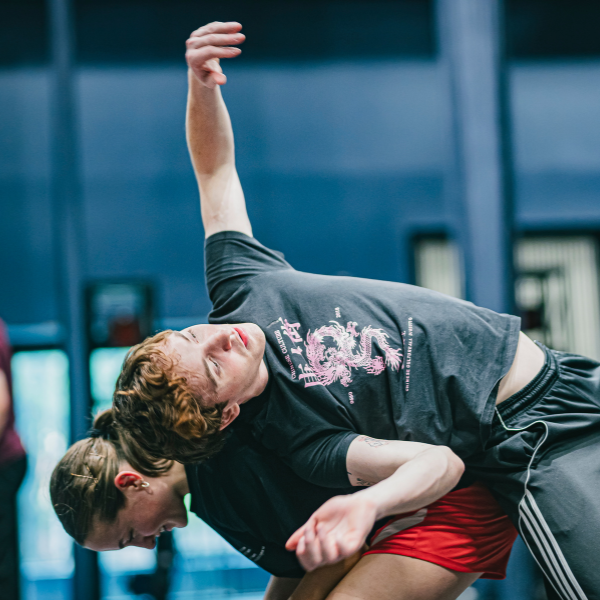 Two young dancers, one leans forwards, the other backwards over the firsts back - arm stretching upwards