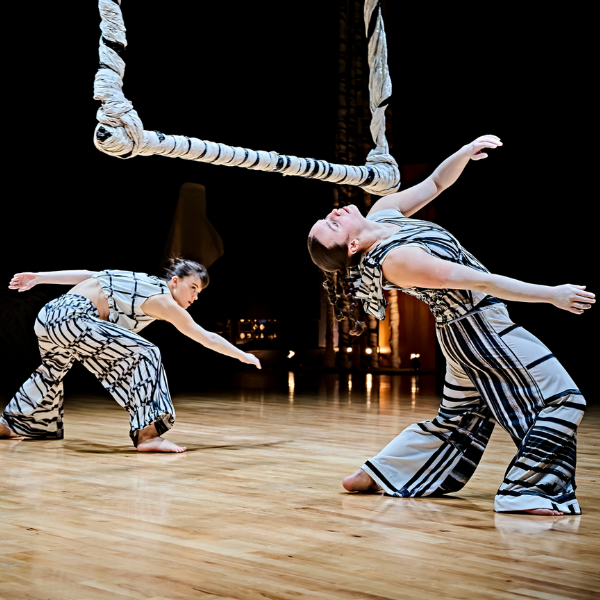 two dancers in white costumes printed with charcoal line drawings bend backwards under a swinging sculpture made of twisted paper that hangs from the roof