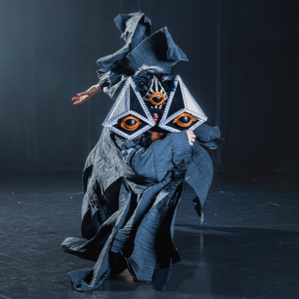 a dancer in a bird mask flings their arm above and behind them, robes flowing and flying as they do so