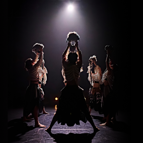 dancers stand in a circle under a bright light, all facing the centre and raising their headpieces aloft