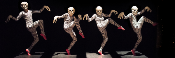 four skeletons creep across the stage in creepy masks and striped leotards, their knees are high and they wear red ballet shoes