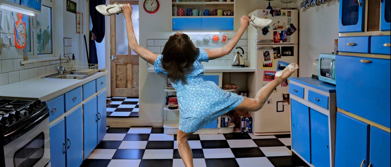 Roots image, dancer in a blue kitchen with back to the camera and legs in the air holding shoes.