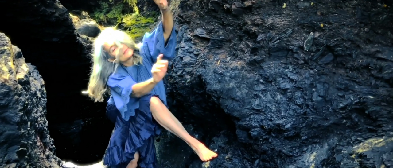 Jo Shapland in a blue dress, dancing barefoot on rocks near the sea