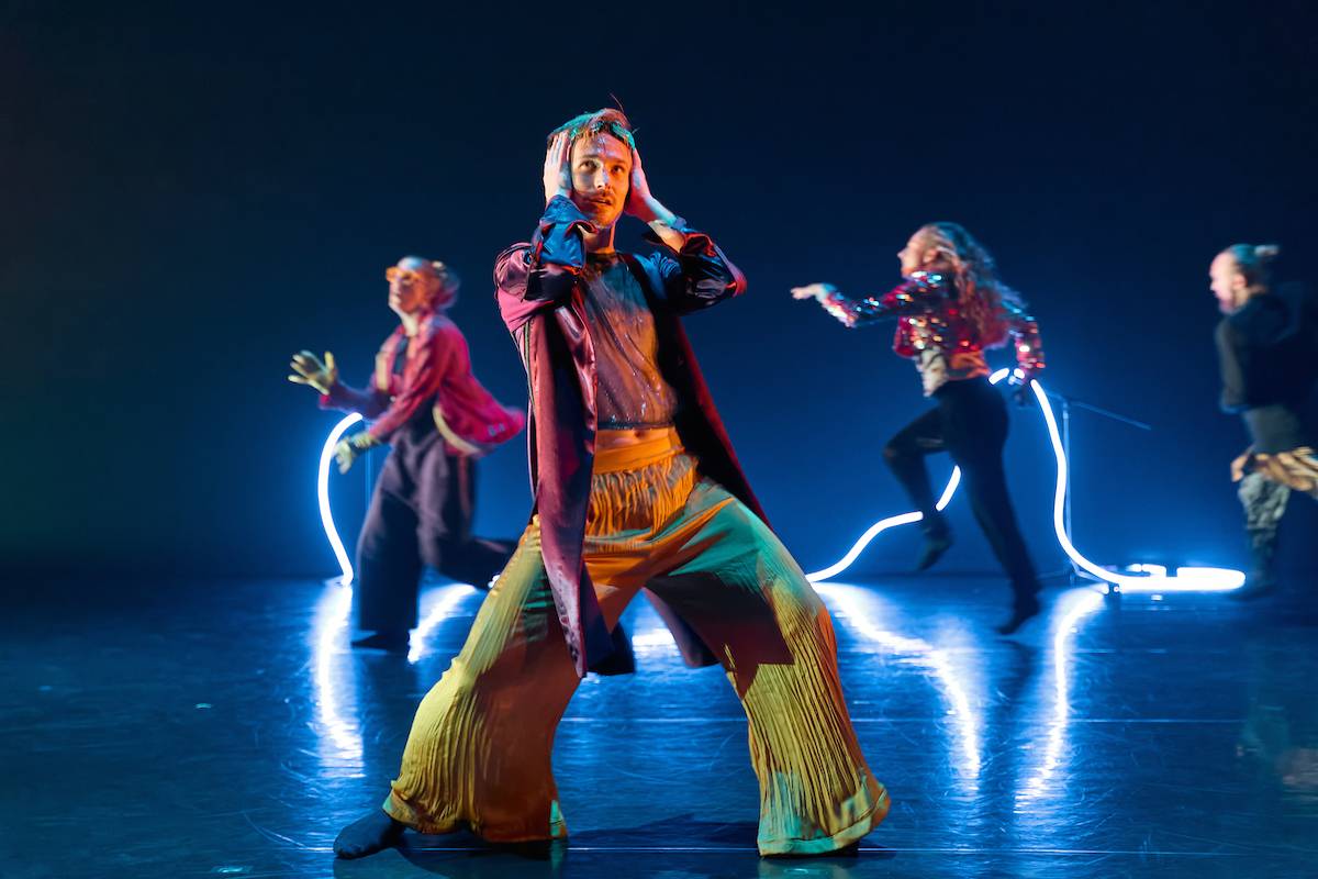 a male dancer in a sparkly jacket and yellow trousers drags his hands through his hair in front of a blue neon light