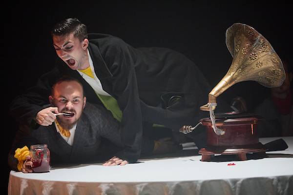 Two dancers in suits and yellow cravats lean over a table, one forces the other to eat jam