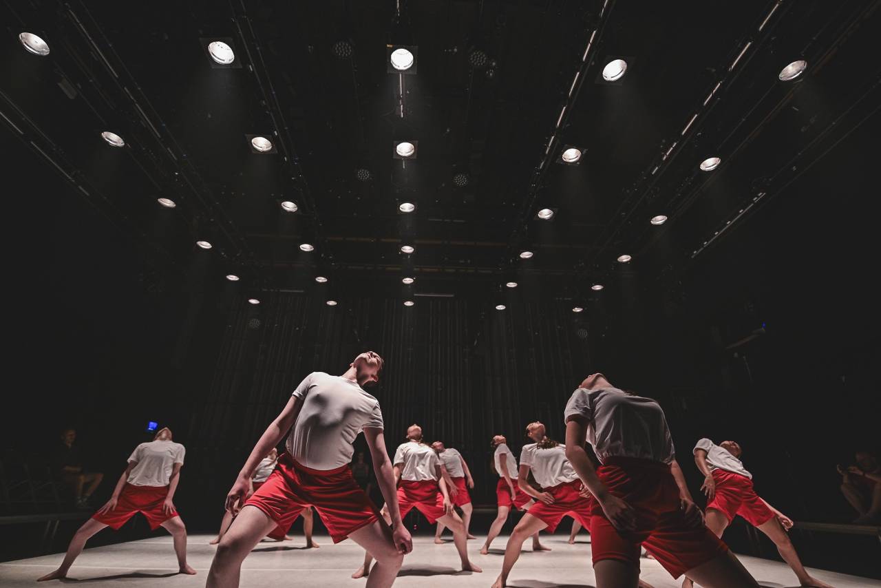 a number of young dancers in white t-shirts and red sports shirts leaning back under rows of spotlights