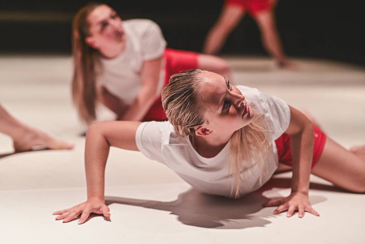 brighly lit young dancer leans across the floor on her knees and hip, holding herself up by the arms and looking to the side