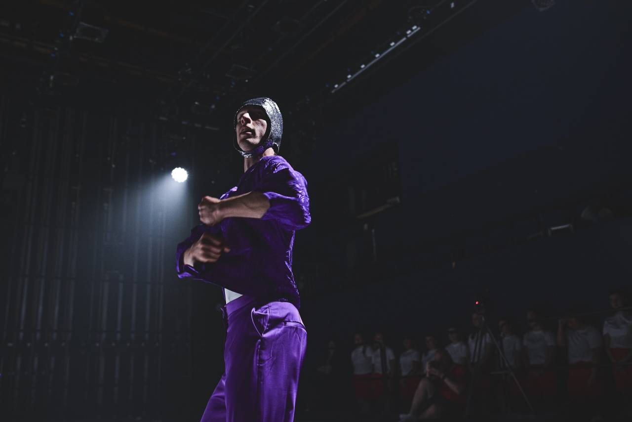 a dancer in a purple satin outfit and dramatic lighting with swim cap holds his hands in fists near his chest
