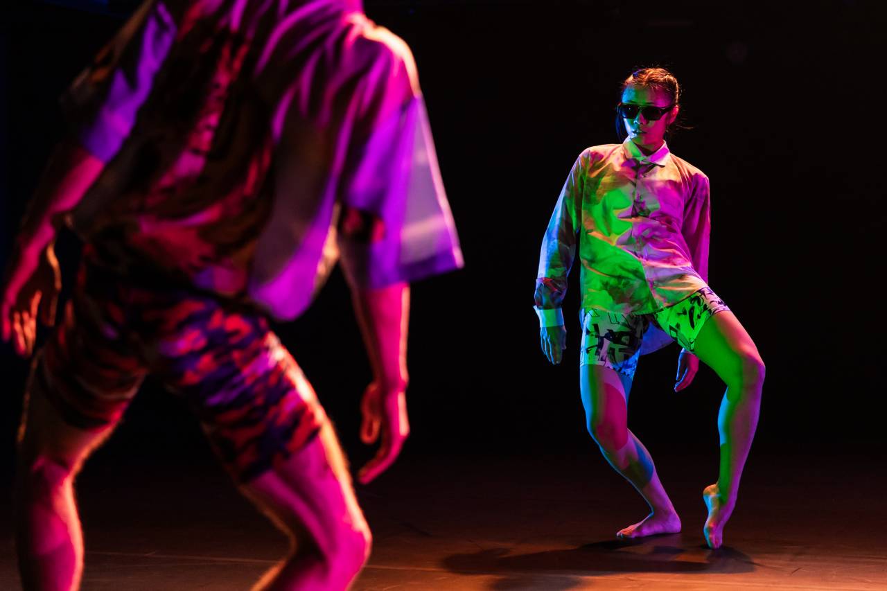 two dancers under neon lights in fuky shirts and shorts, Jill wears sunglasses and faces the camera, one knee up