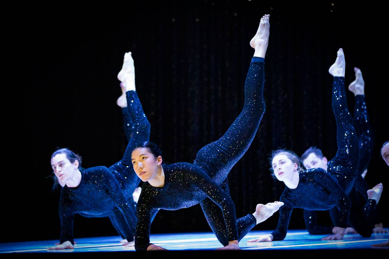 Faye balances on one knee and her hands, one leg pointed towards the sky, she wears a black sequin leotard 