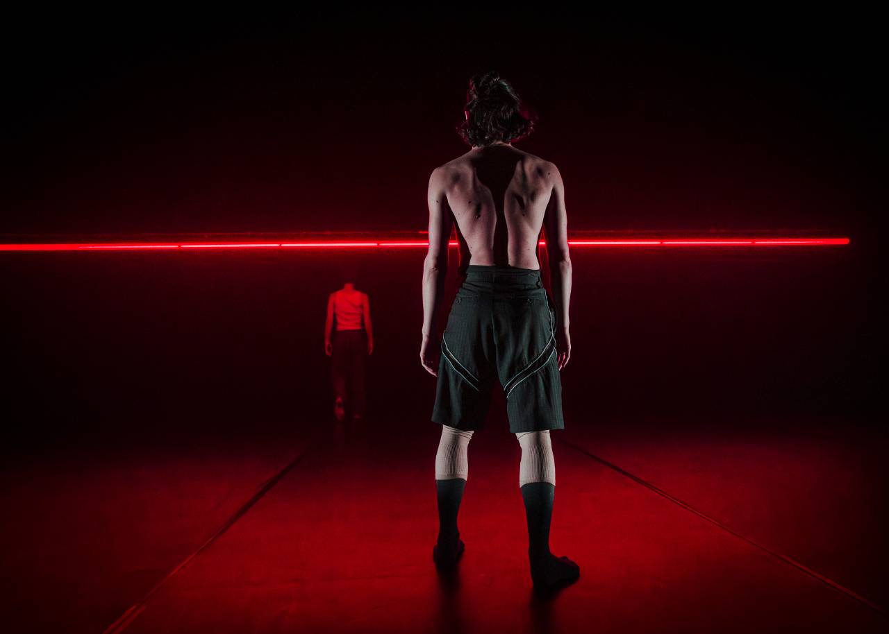 a dancer stands shirtless infront of a red bar of light, looking into the distance 