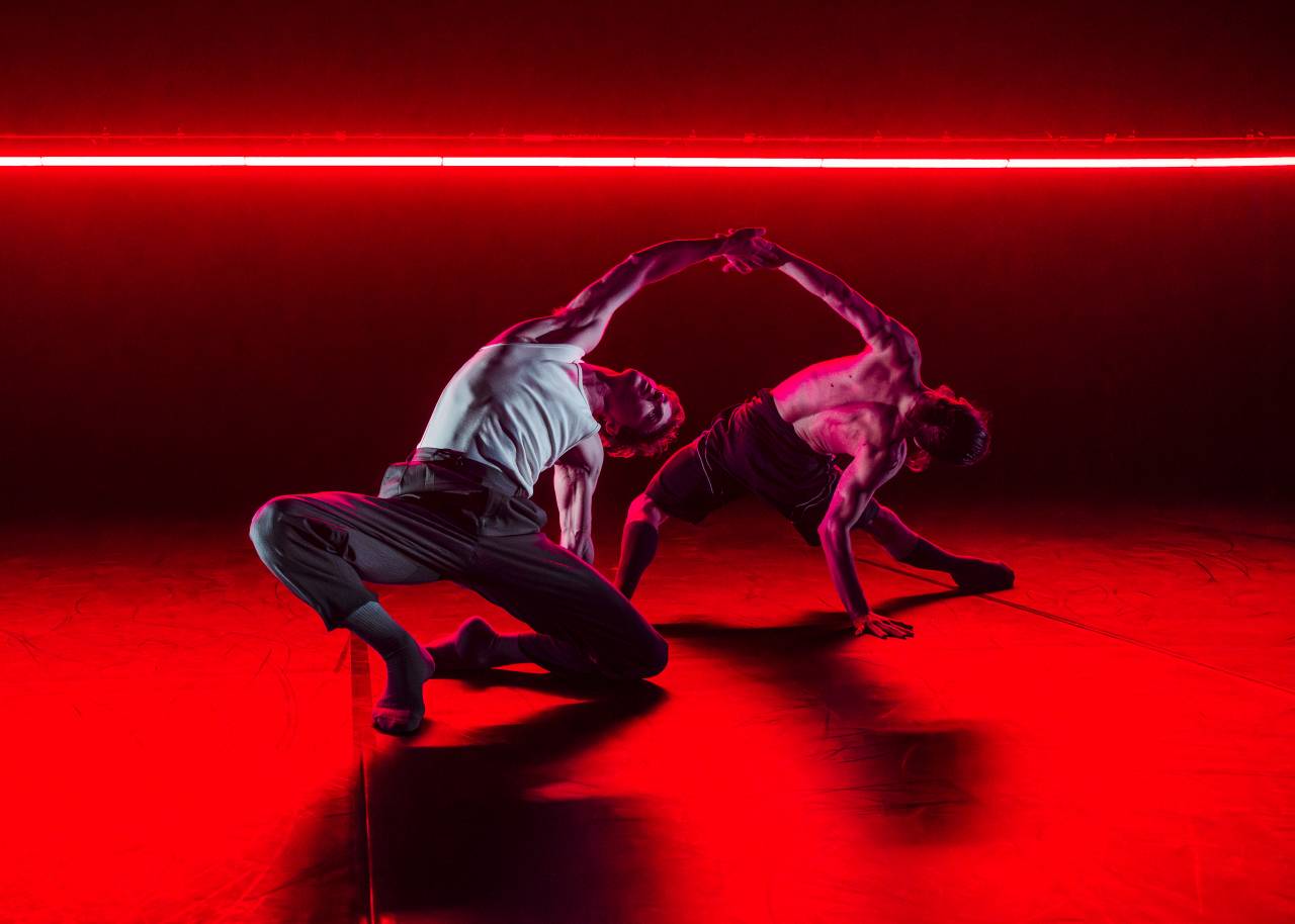 two dancers hold hands as they skim away from one another, feet and hands on the floor under a dramatic red light