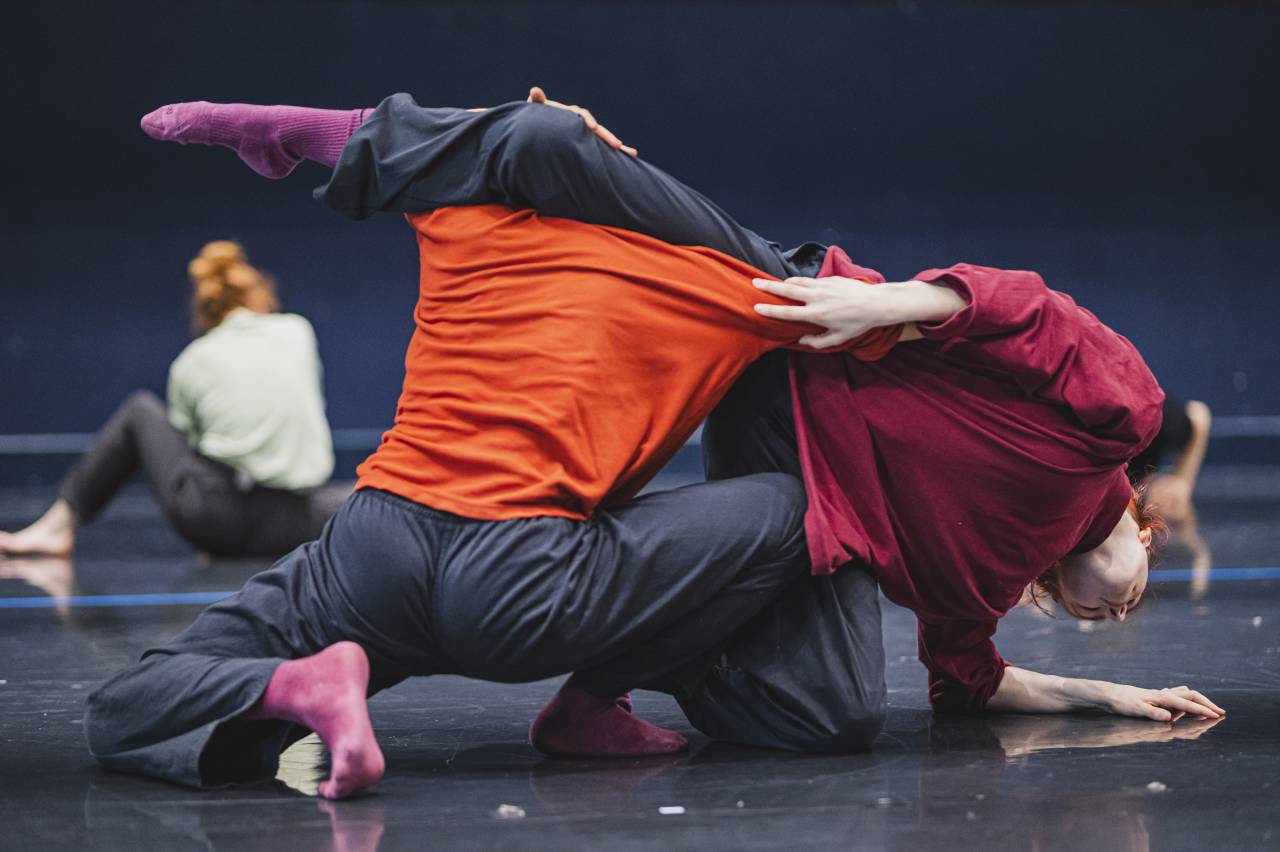 Nimah and Faye bend around one another, Niamh's leg over Faye's shoulders in rehearsal for August