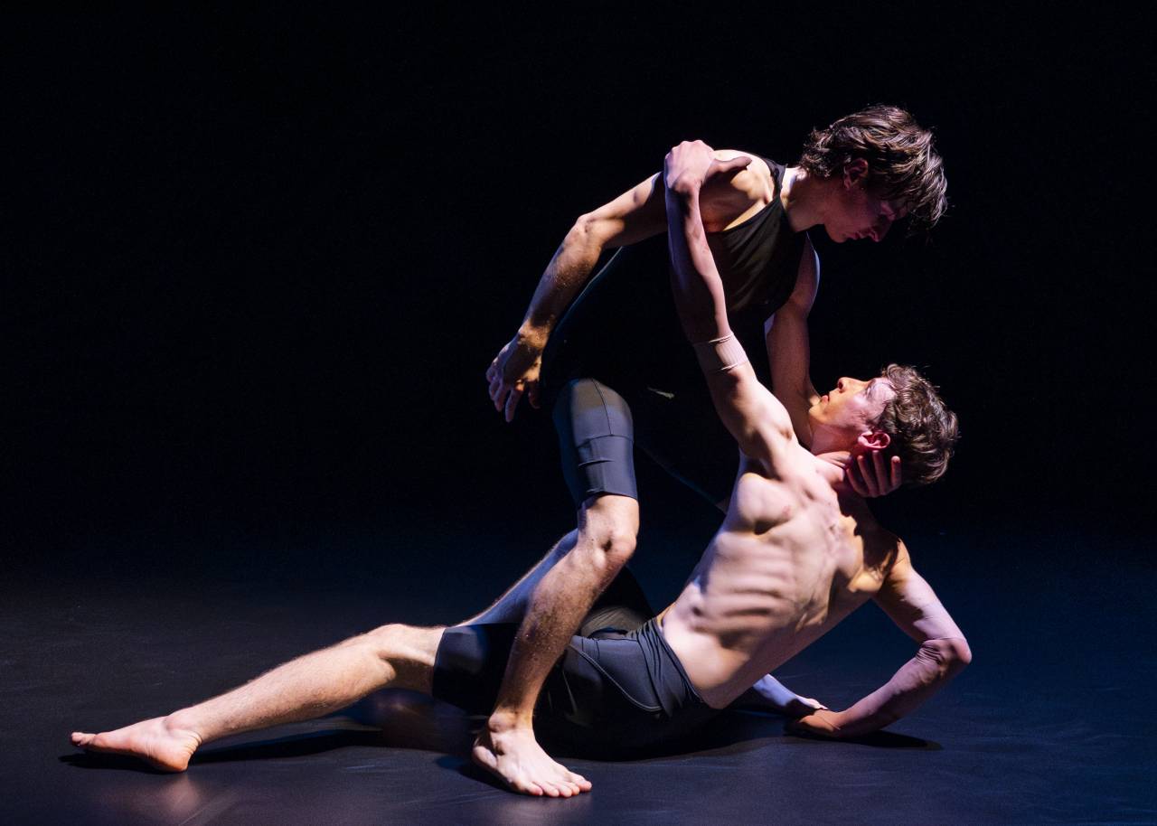 two dancers, one topless holds onto the other as if seeking somehthing