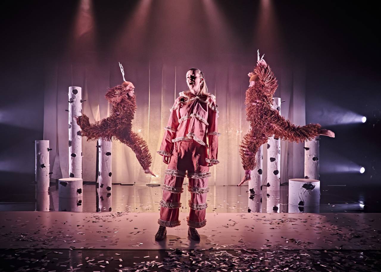 two dancers dressed as deer leap across a sepia lit stage with a wintery set, between them a dancer in a print traditional looking snow outfit with white trim addresses the audience 