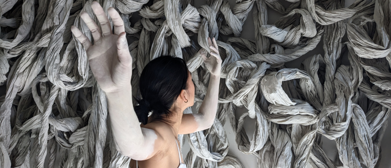 a dancer stands back to the camera, one arm pointing towards the lens, the other away, her arms are painted white and she stands facing a large sculpture made of natural coloured twisted paper