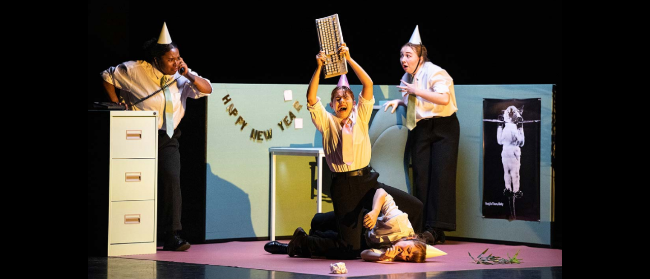 a small set of an office made of two green walls, a filing cabinet and a poster of a cat hanging from a branch that says 'hang in there, baby', a banner on the wall reads 'happy new year' and three dancers in office wear and party hats stand around one lying on the floor as another appears to hit them with a keyboard