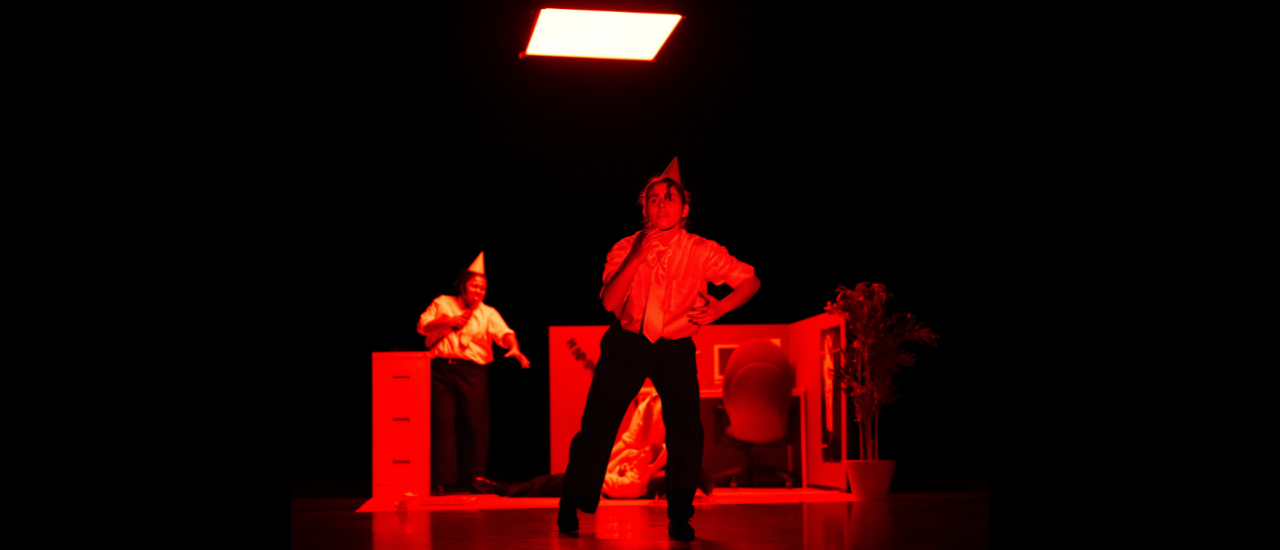 a vibrant red light covers the stage and set whilst a dancer dressed as an office worker and wearing a party hat addresses the audience in front of a small set of an office