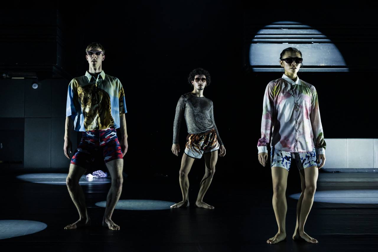 Pietro is at the back of the stage, two others stand downstage of him, they all wear bright streetwear and sunglasses, posing under circles of light in rehearsal for Catachory by Boram Kiim