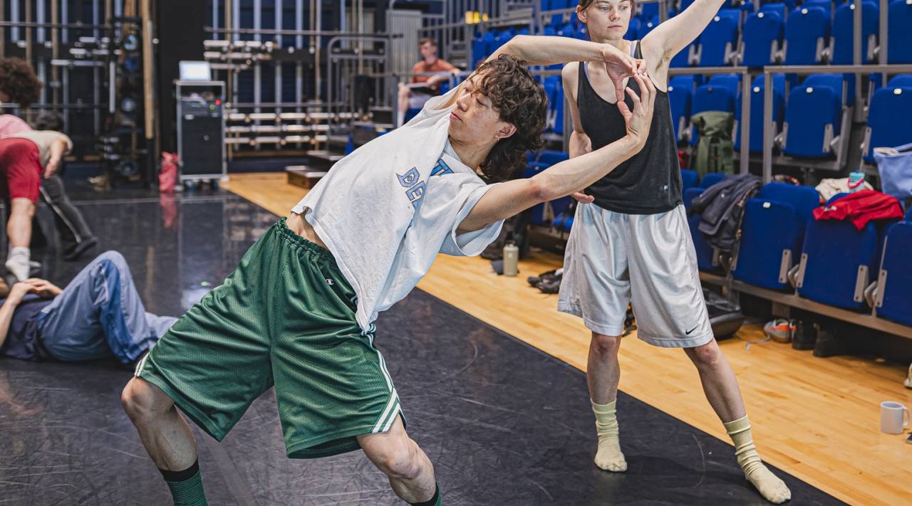 Jason in rehearsal for Mabon in a bright studio, leaning back dramatically arms overhead and wearing green rehearsal shorts and white t shirt
