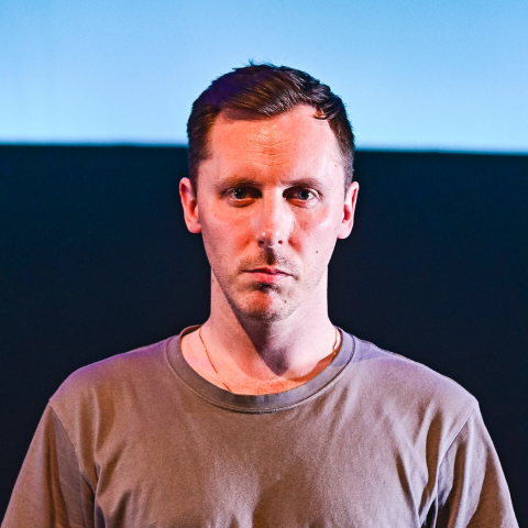 Matthew is a white man with short hair, his photo is bathed in strong, red light.