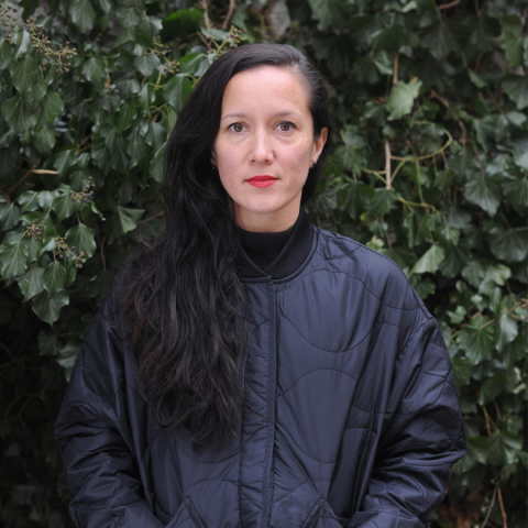 Melanie has long black hair and red lipstick, she stands infront of a lush hedge