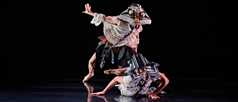 a dancer in a strikign bird mask towers over another on lies on the floor wearing a fish mask - both are mid movement