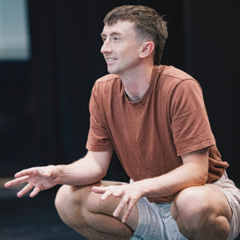 Osian is crouched in a dance studio smiling as they direct the dancers of screen. They wear a rust coloured t shirt and grey shorts.