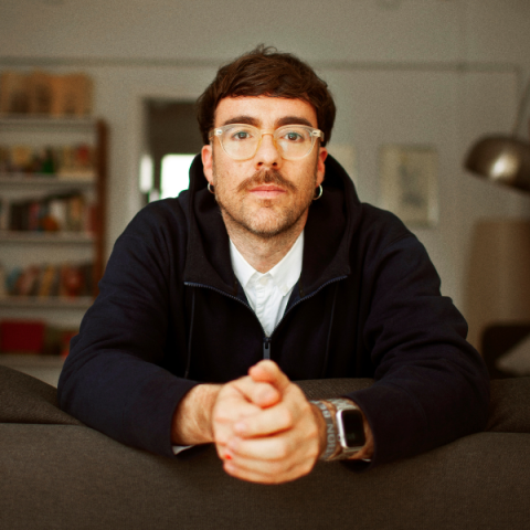Marcos Morau sits on the floor in front of a trendy book case, he is white with a moustache and wears glasses