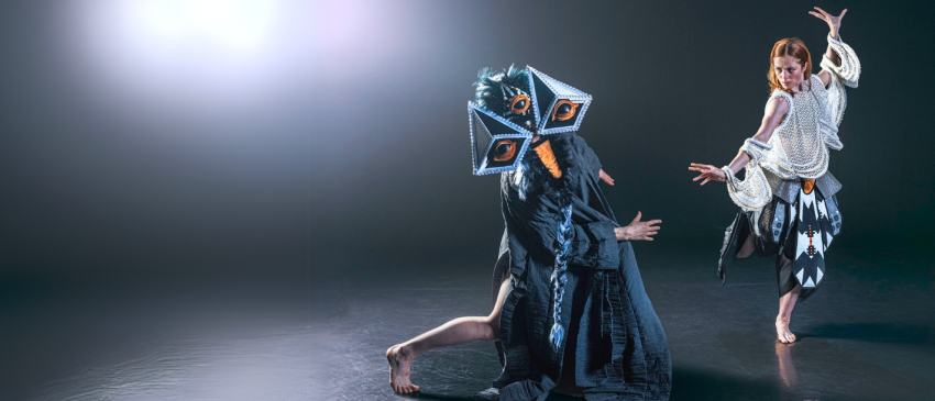 two dancers dressed in wild costumes, one in a large folk inspired bird mask and the other in a top that looks inspired by skeletons, they pose dynamically under blooming light